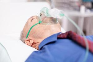 Senior man getting treatment for lungs infected with covid19 wearing oxygen mask. Patient in hospital room laying on medical bed with infected lung during coronavirus pandemic having respiratory problem. photo