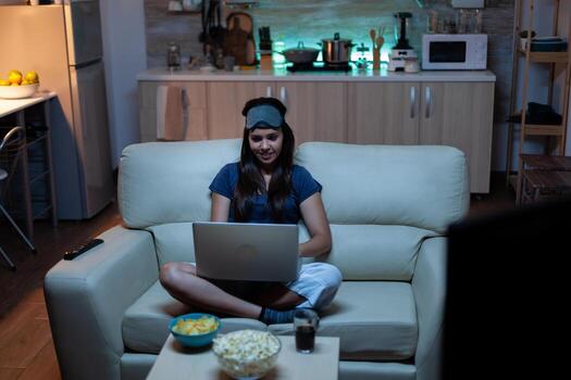 Lady in pijamas and eye cover on forehead typing on laptop and watching tv late at night . Freelancer working sitting on sofa reading writing searching browsing on notebook using internet technology photo