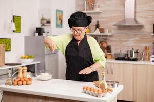 retirado cocinero preparando Pizza en hogar cocina vistiendo delantal. contento mayor cocinero con uniforme aspersión, tamizado cernido crudo ingredientes por mano en hogar cocina. foto