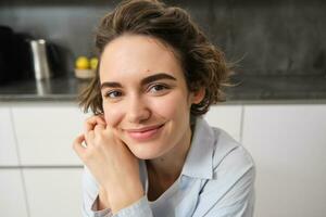 cerca arriba retrato de joven morena mujer, mira a cámara y sonrisas, se sienta a hogar en cocina foto