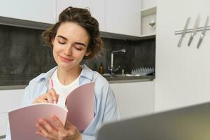 Young woman checking her schedule while working on remote from home, using laptop, looking at her daily planner, making notes, writing down information in notebook photo