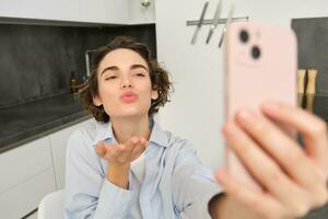 Portrait of beautiful young woman, taking selfie on mobile phone at home, poses for photo with smartphone