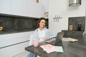 Portrait of young beautiful woman, studies at home, flips her workbook, works from home, uses laptop to learn on remote, makes notes during webinar photo