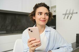 Image of young modern woman, holding mobile phone, sitting in her kitchen at home, order delivery on smartphone app photo