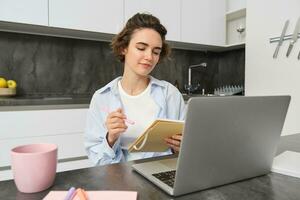 Working woman makes notes, writes down information on notebook, sitting with laptop at home photo