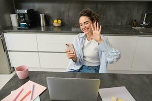 Young friendly woman, waves hand at laptop camera, video chats, holds smartphone, has conversation online photo