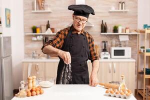 Retaried baker wearing apron and prepraing homemade pizza on kitchen table. Retired senior chef with bonete and apron, in kitchen uniform sprinkling sieving sifting ingredients by hand. photo