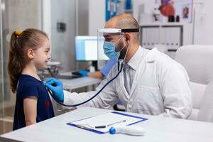 Pediatrician doctor examining little girl with stethoscope for disease in the medical office. Physician specialist in pediatry medicine providing professional treatment services in protective equipment photo