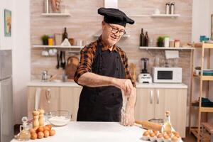 Smiling experienced male baker making homemade cookies wearing apron and bonete. Retired senior chef in kitchen uniform sprinkling sieving sifting ingredients by hand. photo
