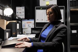 Accountancy asian employee clocking out from work, shutting down laptop in business office. Bookkeeper executive in archival depository filled with cabinet, folders and flowcharts photo