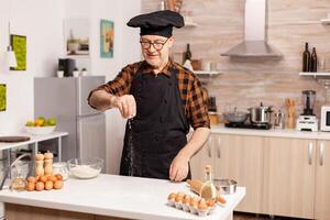 Experienced baker in kitchen preparing delicious pizza using bio wheat flour. Retired senior chef with bonete and apron, in kitchen uniform sprinkling sieving sifting ingredients by hand. photo