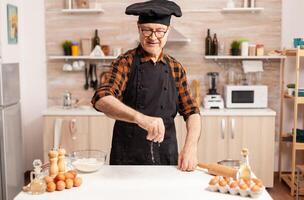 Retired cook preparing tasty food spreading wheat flour on kitchen table top. Retired senior chef with bonete and apron, in kitchen uniform sprinkling sieving sifting ingredients by hand. photo