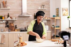 Senior woman recording live video for culinary vlog at home. Retired blogger chef influencer using internet technology communicating, shooting blogging on social media with digital equipment photo