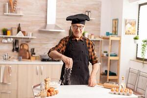 Elderly baker preparing tasty food using bio wheat flour wearing apron and bonete. Retired senior chef with bonete and apron, in kitchen uniform sprinkling sieving sifting ingredients by hand. photo
