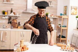 Retired chef in home kitchen wearing apron while sprinkling ingredient on table for tasty. Retired senior chef with bonete and apron, in kitchen uniform sprinkling sieving sifting ingredients by hand. photo