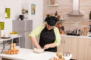 mayor mujer vistiendo bonete mientras preparando masa para pegar en hogar cocina siguiendo tradicional receta. retirado mayor cocinero con uniforme aspersión, tamizado cernido crudo ingredientes y mezclando foto