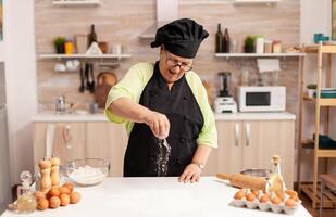 Portrait of cheerful senior lady spreading flour on kitchen table during cookies preparation. Happy elderly chef with uniform sprinkling, sieving sifting raw ingredients by hand. photo