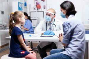 Physician having conversation with child during consultation and coronavirus outbreak in hospital office wearing face mask. Health pediatrician specialist providing health care services consultations treatment. photo