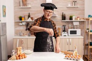 Senior baker sprankling wheat flour to prepare delicious cookies wearing bonete. Retired chef with bonete and apron, in kitchen uniform sprinkling sieving sifting ingredients by hand. photo