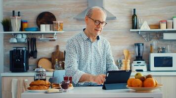 Senior parent browsing on tablet pc in kitchen during breakfast having a healthy lifestyle. Elderly person with tablet portable pad PC in retirement age using mobile apps, modern internet online information technology with touchscreen photo