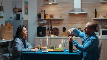 Young enthusiastic man opening a bottle of red wine during romantic dinner. Happy couple talking, sitting at table in kitchen, enjoying the meal, celebrating their anniversary at home with healty food photo