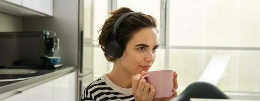 cerca arriba retrato de sonriente morena mujer, estudiante bebidas su té y escucha música o libro electronico en auriculares, se sienta en cocina y relaja foto