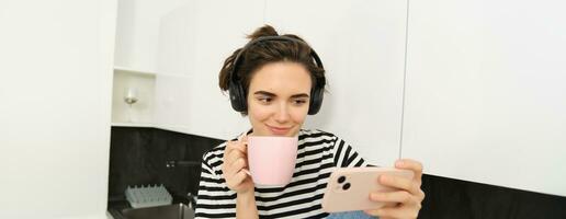 Smiling young woman watching tv series or video blog on smartphone app, drinking tea in the kitchen, looking at mobile phone with interest photo