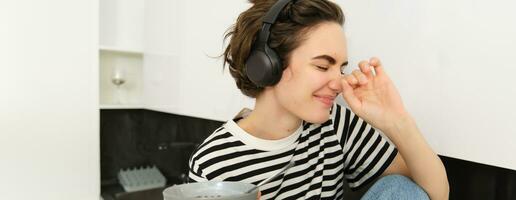 Portrait of carefree woman in headphones, eating her breakfast, holding bowl of cereals, laughing and smiling, sitting in the kitchen on a counter photo