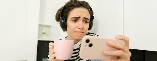 cerca arriba retrato de joven mujer en auriculares, acecho alguna cosa triste en teléfono inteligente solicitud, ceñudo y mirando con simpatía a móvil teléfono pantalla, Bebiendo té o café en el cocina foto