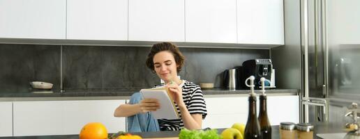 Portrait of young smiling woman in kitchen, holding notebook, making notes for recipe, writing grocery list, cooking salad, sitting near vegetables and chopping board photo