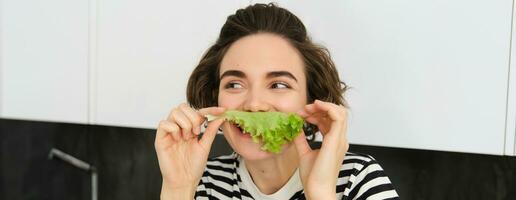 Cute smiling woman eating lettuce leaf and smiling, having a healthy snack, likes vegetables, posing in the kitchen photo