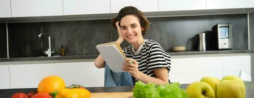 Portrait of young smiling woman in kitchen, holding notebook, making notes for recipe, writing grocery list, cooking salad, sitting near vegetables and chopping board photo