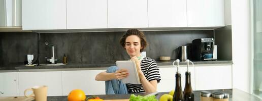 Portrait of young smiling woman cooking with vegetables, writing down ingredients, looking at her notebook, reading recipe, sitting in kitchen photo