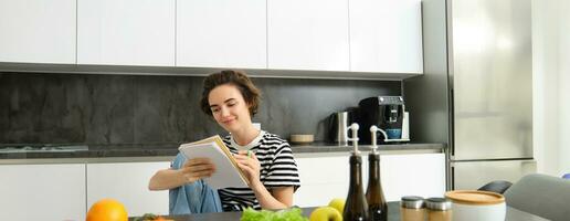 Portrait of young woman cooking, writing notes, grocery list in notebook, creating list of meals to cook through meal, sitting in kitchen near vegetables and chopping board photo
