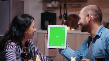 Cheerful young couple using green mockup screen digital isolated tablet computer. Husband and wife looking at green screen template chroma key display sitting at the table in kitchen during dinner. photo