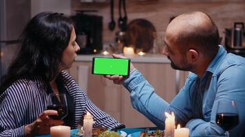 Married young couple holding mockup phone at dinner. Happy looking at green screen template chroma key isolated smart phone display using techology internet sitting at the table in kitchen. photo