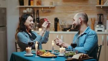 Handsome man proposing to his girlfriend marriage during festive dinner, in kitchen sitting at the table drinking a glass of red wine. Happy surprised woman smiling and hugging him. photo