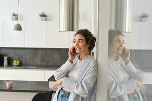 Beautiful woman talking on mobile phone, answers phone call and smiles, stands at home, leans to wall and chats photo