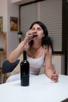 Alcoholic woman having bottle of wine and glass feeling sad at home. Lonely person drinking beverage with alcohol being depressive. Adult with addiction feeling emotional and upset photo