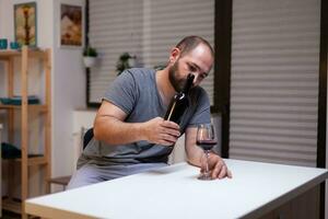 Lonely man pouring wine in glass from bottle drinking alone feeling depressed and unhealthy. Miserable adult with alcohol addiction being intoxicated with liquor, booze, beverage photo