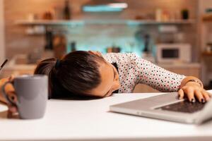 Freelance woman falling asleep in home kitchen working overtime on a project. Employee using modern technology at midnight doing overtime for job, business, busy, career, network, lifestyle ,wireless. photo