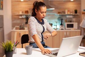 Woman checking watch during overtime work in home kitchen at midnight. Employee using modern technology at night hours doing overtime for job, business, busy, career, network, lifestyle ,wireless. photo