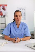 retrato de bonito médico enfermero sonriente a cámara en hospital oficina vistiendo azul uniforme. cuidado de la salud facultativo sentado a escritorio utilizando computadora en moderno clínica mirando a monitor, medicamento. foto