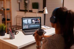 Videographer holding cup of coffee while working in video footage during post production. Content creator in home working on montage of film using modern software for editing late at night. photo