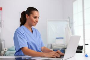 Medical nurse in uniform using laptop sitting at desk in hospital office. Health care physician using computer in modern clinic looking at monitor, medicine, profession, scrubs. photo