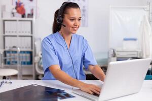 Medical receptionist wearing headset with microphone in private hospital typing on laptop Health care physician sitting at desk using computer in modern clinic looking at monitor. photo
