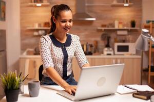 concentrado mujer de negocios sonriente trabajando tarde a noche utilizando ordenador portátil en cocina. empleado utilizando moderno tecnología a medianoche haciendo tiempo extraordinario para trabajo, negocio, ocupado, carrera profesional, red, estilo de vida. foto