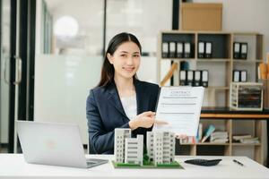 Young real estate agent worker working with laptop and tablet at table in office and small house beside it. photo