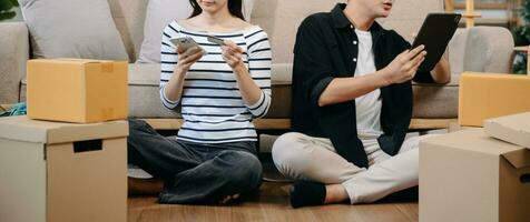 asiático joven atractivo Pareja hombre y mujer utilizar tableta y teléfono inteligente en línea compras mueble Decorar casa con caja de cartón paquete moverse en nuevo casa. foto