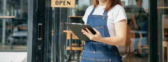 Young female hanging a welcome sign in front of a coffee shop. Beautiful waitress or hostess holding a tablet preparing  in a restaurant. photo